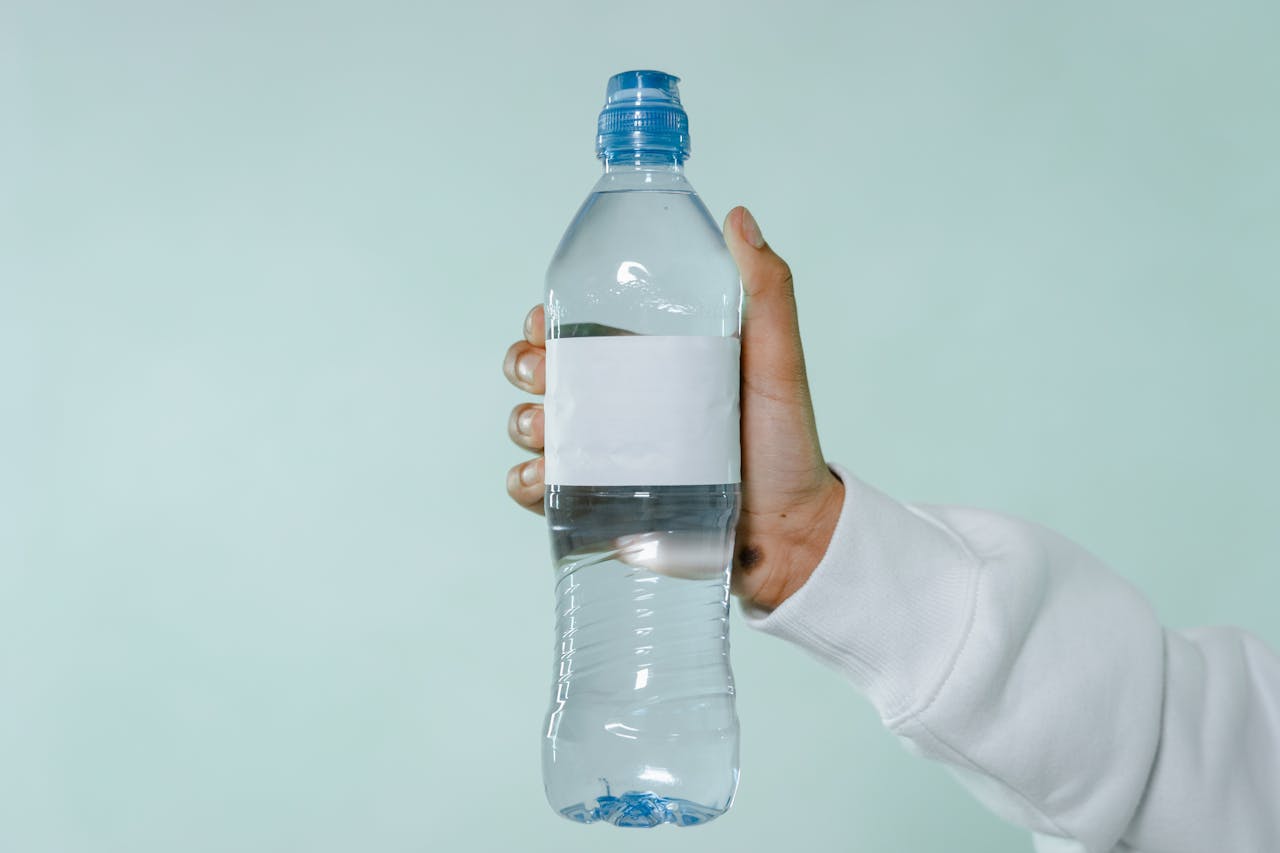 A hand holding a clear plastic water bottle with blank label on a soft green background.